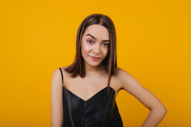 Chica interesada con cabello castaño oscuro posando. Encantadora mujer caucásica joven en camiseta negra de pie delante de la pared amarilla.