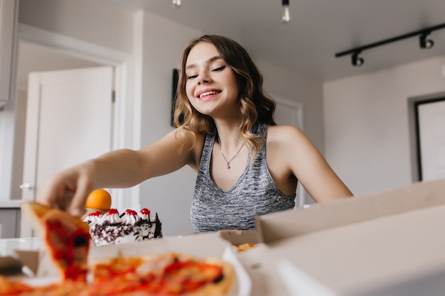 Foto gratuita chica interesada con peinado ondulado comiendo pizza con placer. modelo de mujer glamorosa sentada en la cocina y disfrutando de la comida rápida.