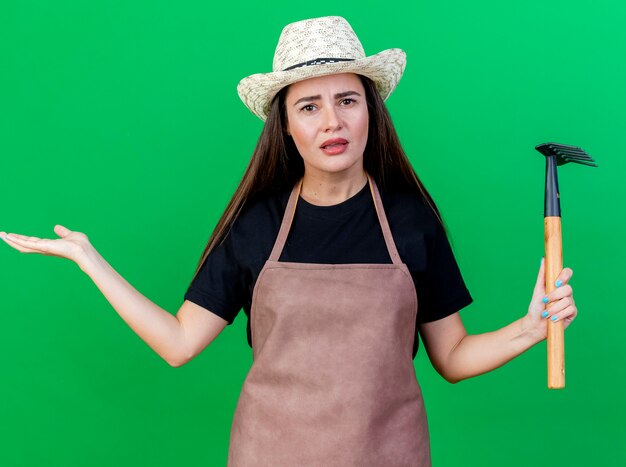 Chica jardinero hermosa confundida en uniforme con sombrero de jardinería sosteniendo rastrillo mano extendida aislada en verde