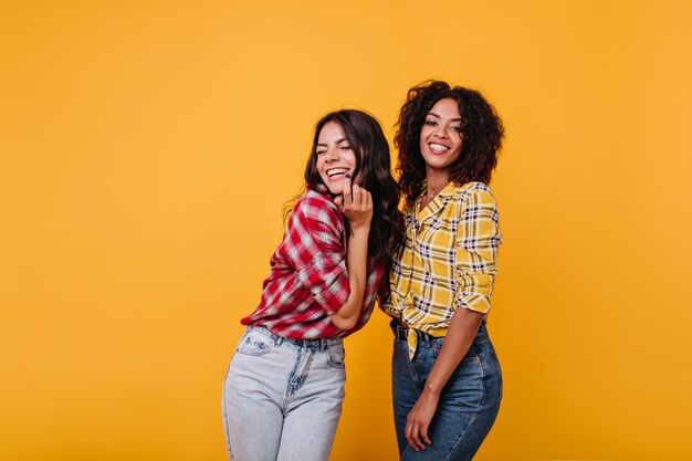 Chica en jeans claros se ríe de todo corazón con las bromas de su novia mulata. Instantánea en interiores de mujeres de buen humor.