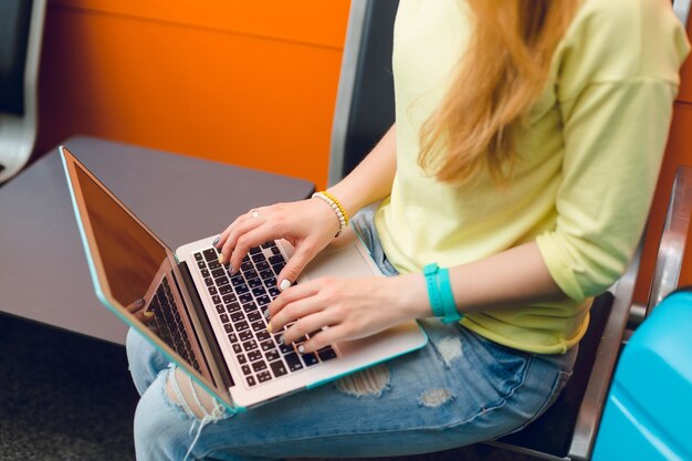 Chica en jeans y suéter amarillo está sentada en una silla. Ella tiene una computadora portátil en las rodillas. Concéntrese en escribir a mano en la computadora portátil.