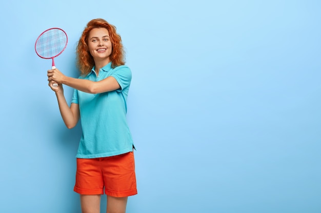 Chica de jengibre activa sostiene una raqueta de tenis, vestida con una camiseta azul informal y pantalones cortos rojos, disfruta del juego con un amigo, tiene una expresión feliz