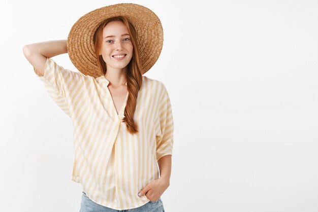 Chica de jengibre atractiva elegante y alegre con pecas sosteniendo la mano detrás de la tapa de paja y sonriendo ampliamente teniendo una gran mañana caminando por la playa