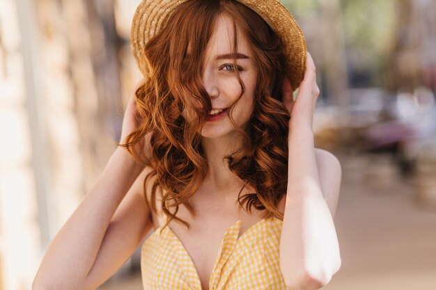 Chica de jengibre de buen humor en ropa amarilla posando en la calle borrosa. Refinada mujer pelirroja con sombrero pasar tiempo en la ciudad.