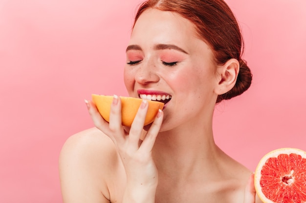 Chica de jengibre complacida comiendo pomelo. Disparo de estudio de mujer sensual disfrutando de frutas sobre fondo rosa.