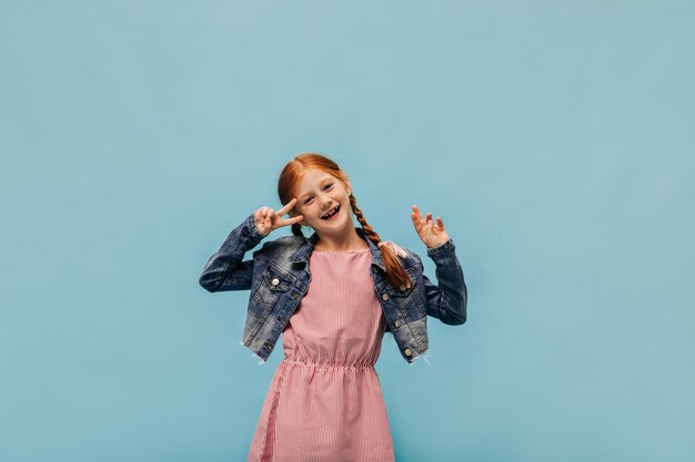 Chica de jengibre de moda con pecas en chaqueta y vestido moderno rosa mostrando el signo de la paz y sonriendo en la pared aislada