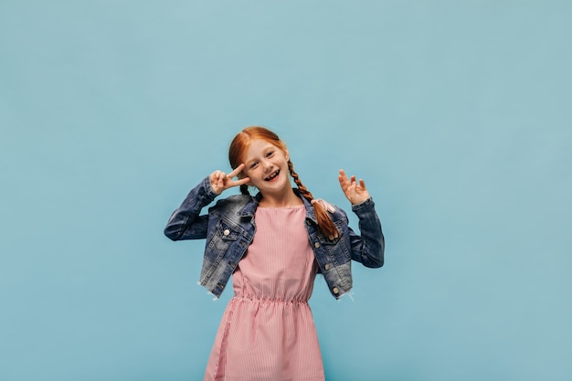 Chica de jengibre de moda con pecas en chaqueta y vestido moderno rosa mostrando el signo de la paz y sonriendo en la pared aislada