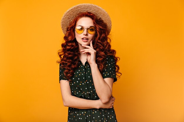 Chica de jengibre pensativa posando en gafas de sol y sombrero de paja. Vista frontal de la romántica dama europea aislada sobre fondo amarillo.