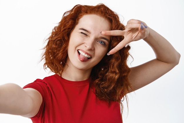 Foto gratuita chica de jengibre positiva con peinado rizado que muestra la lengua y el vsign sobre el ojo tomando selfie o grabando video en la cámara frontal del teléfono inteligente de pie contra el fondo blanco