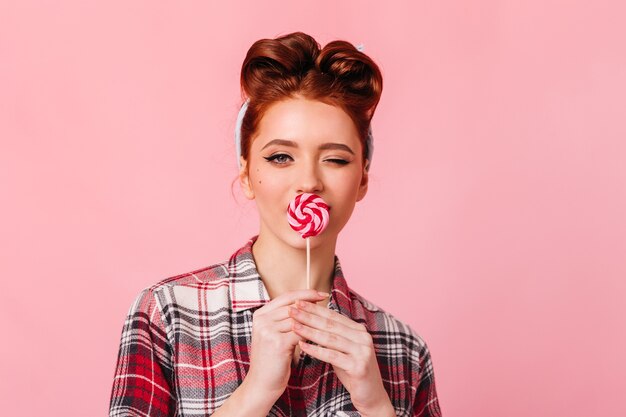 Chica de jengibre sensual comiendo dulces rojos. Vista frontal de la elegante mujer pinup con piruleta aislado en el espacio rosa.