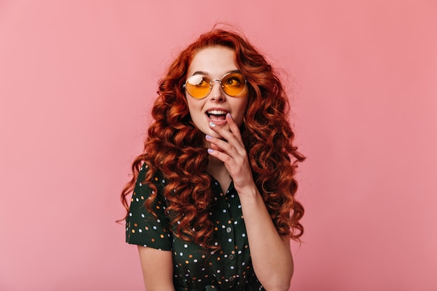 Chica de jengibre soñadora mirando a otro lado con la boca abierta. Disparo de estudio de mujer joven emocional en gafas de sol posando sobre fondo rosa.