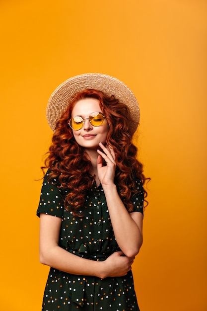 Chica de jengibre soñadora posando con los ojos cerrados. Foto de estudio de increíble mujer caucásica en sombrero de paja aislado sobre fondo amarillo.