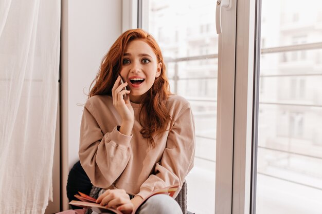 Chica de jengibre sorprendida hablando por teléfono mientras está sentado junto a la ventana. Retrato interior de mujer joven relajada con libro.