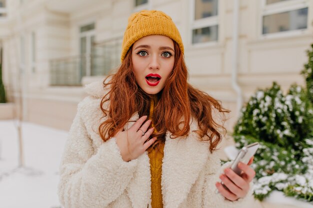 Chica de jengibre sorprendida de pie en la calle en un día frío. Magnífica mujer joven con teléfono posando al aire libre.