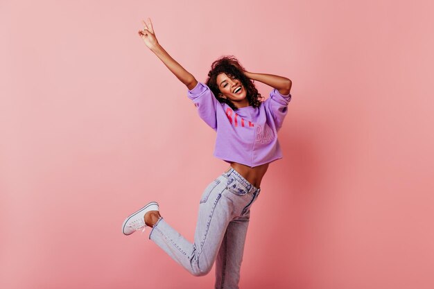 Chica jocund bien formada bailando sobre fondo rosado Dama africana de buen humor saltando con una sonrisa