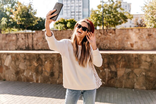 Chica jocund en suéter blanco haciendo selfie en la calle. Mujer bien vestida con boina divirtiéndose en un buen día de otoño.