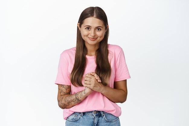 Chica joven de 25 años que parece esperanzada, sonriendo y cogidos de la mano juntos en el pecho, anticipando algo, de pie sobre blanco.