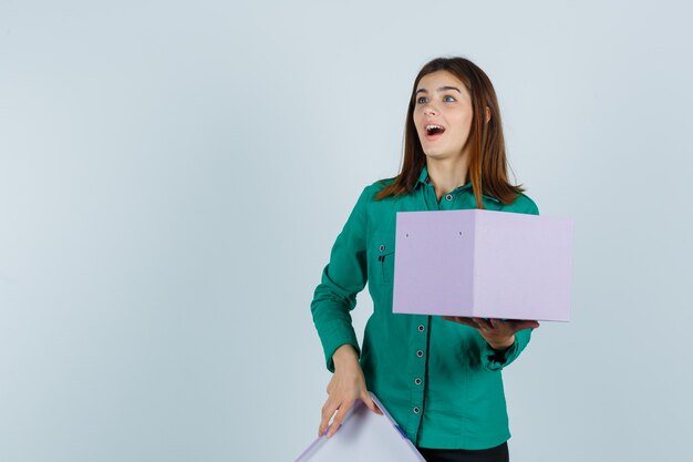 Chica joven abriendo caja de regalo, mirando a otro lado en blusa verde, pantalón negro y mirando sorprendido, vista frontal.