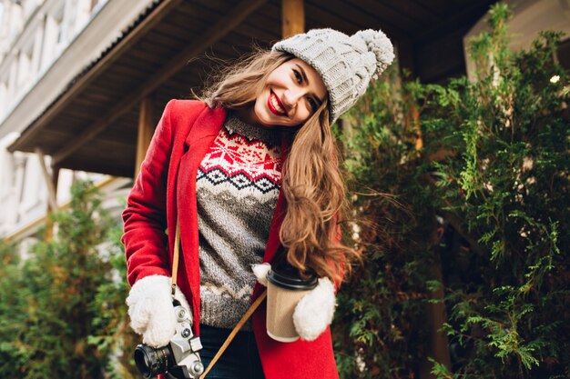 Chica joven en abrigo rojo caminando por la calle con café para llevar. Lleva guantes blancos, sonriendo.