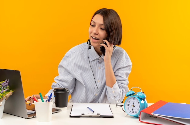 Foto gratuita chica joven alegre del centro de llamadas con auriculares sentados en el escritorio hablando por teléfono y usando la computadora portátil aislado en naranja