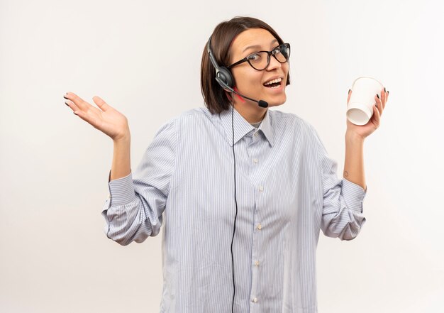 Chica joven alegre del centro de llamadas con gafas y auriculares sosteniendo la taza de café mostrando la mano vacía aislada en blanco