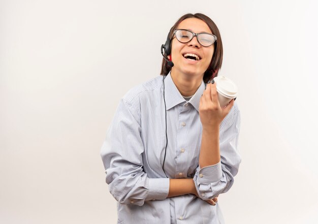 Chica joven alegre del centro de llamadas con gafas y auriculares sosteniendo una taza de café de plástico con los ojos cerrados aislado en blanco