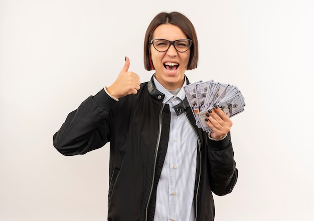 Chica joven alegre del centro de llamadas con gafas sosteniendo dinero y mostrando el pulgar hacia arriba aislado en blanco