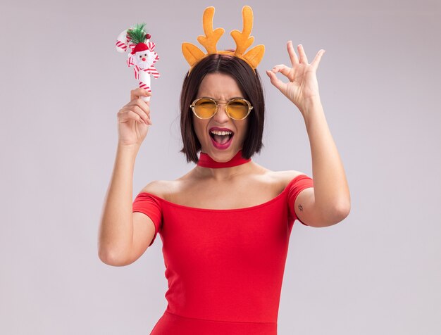 Chica joven alegre con diadema de cuernos de reno y gafas levantando adorno de bastón de caramelo de Navidad mirando a la cámara haciendo bien signo aislado sobre fondo blanco.