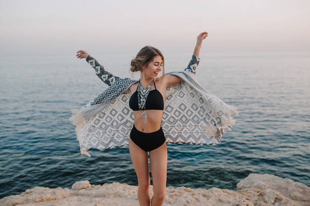 Chica joven alegre que se divierte al atardecer en la playa con rocas, buen tiempo de vacaciones. El uso de elegantes gafas de sol, traje de baño negro de moda, bikini, chaqueta de punto, capa con adornos.