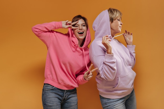 Chica joven alegre en sudadera rosa mostrando signos de paz, guiñando un ojo y posando con una mujer moderna en sudaderas con capucha lila sobre fondo naranja.