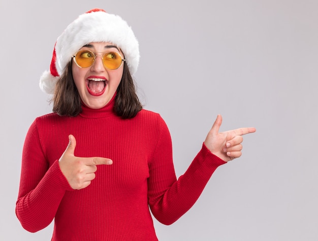 Chica joven alegre en suéter rojo y gorro de Papá Noel con gafas mirando a un lado apuntando con los dedos índices al lado de pie sobre la pared blanca