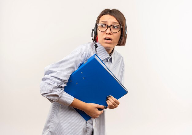 Foto gratuita chica joven ansiosa del centro de llamadas con gafas y auriculares con carpeta mirando al lado aislado en blanco con espacio de copia