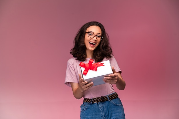 Foto gratuita chica joven en anteojos ofreciendo una caja de regalo blanca y sonriendo.