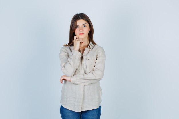 Chica joven apoyando la barbilla en la mano con camisa beige, jeans y mirando serio, vista frontal.