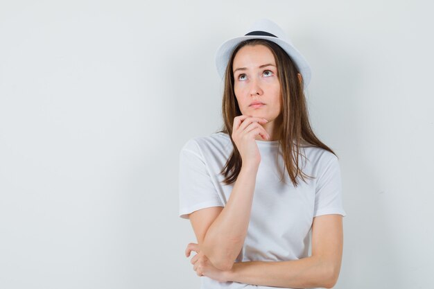 Chica joven apoyando la barbilla en la mano con camiseta blanca, sombrero y aspecto vacilante. vista frontal.