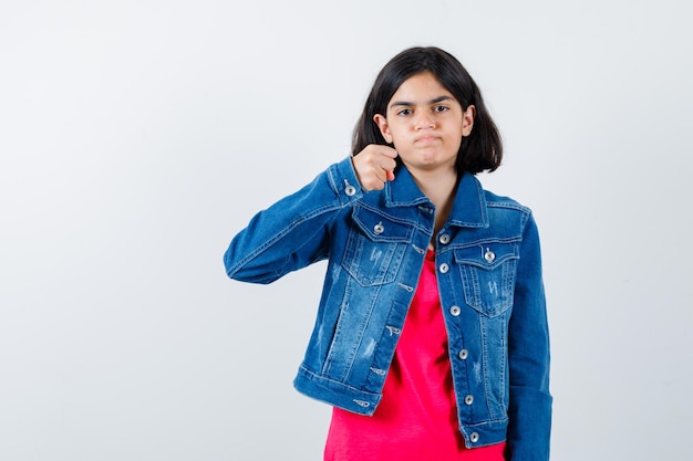 Chica joven apretando el puño en camiseta roja y chaqueta de jean y mirando seria. vista frontal.