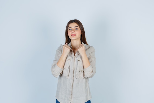 Foto gratuita chica joven apretando los puños sobre el pecho en camisa beige, jeans y mirando alegre. vista frontal.