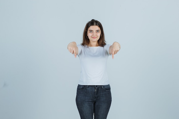 Foto gratuita chica joven apuntando hacia abajo en camiseta, jeans y mirando confiado, vista frontal.