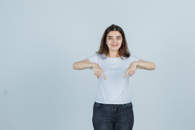 Chica joven apuntando hacia abajo en camiseta, jeans y mirando confiado, vista frontal.