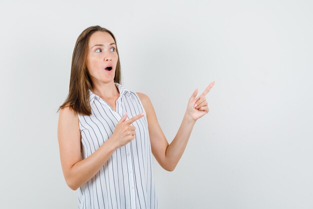 Chica joven apuntando hacia afuera en camiseta y mirando curiosa, vista frontal.