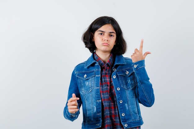 Chica joven apuntando hacia arriba en camisa a cuadros y chaqueta de jean y mirando serio