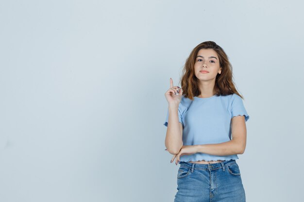 Chica joven apuntando hacia arriba en camiseta, jeans y mirando confiada. vista frontal.