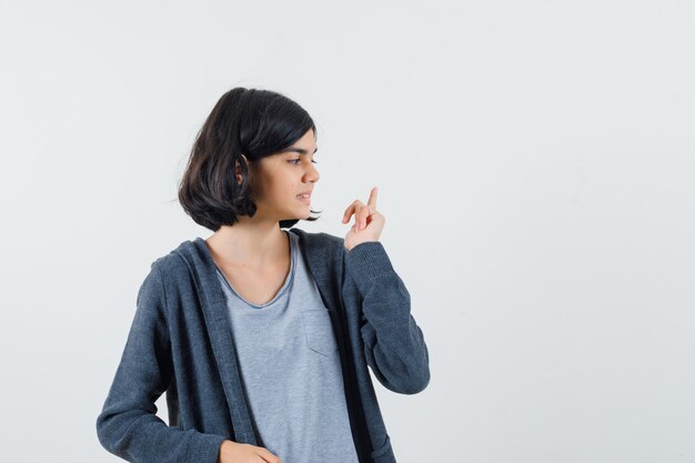 Chica joven apuntando hacia arriba con el dedo índice mientras mira hacia otro lado con una camiseta gris claro y una sudadera con capucha gris oscuro con cremallera y se ve linda