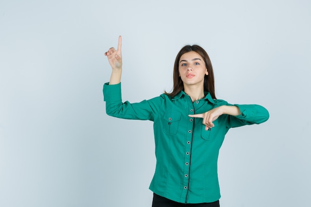 Foto gratuita chica joven apuntando hacia arriba y hacia la izquierda con el dedo índice en blusa verde, pantalón negro y mirada seria. vista frontal.