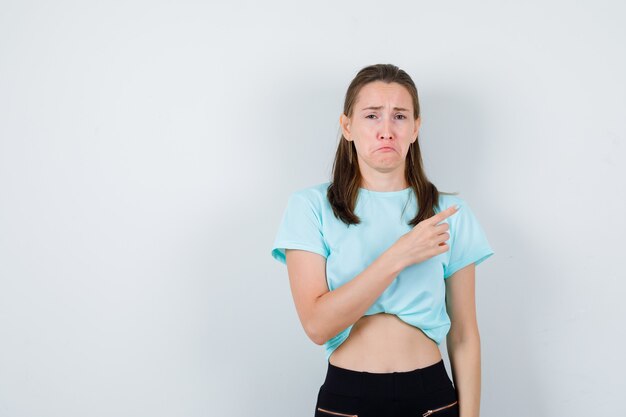 Chica joven apuntando hacia arriba, labios curvados en camiseta turquesa, pantalones y mirando agraviado, vista frontal.