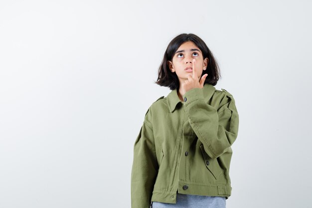 Chica joven apuntando hacia arriba con suéter gris, chaqueta caqui, pantalón de mezclilla y mirando enfocado, vista frontal.