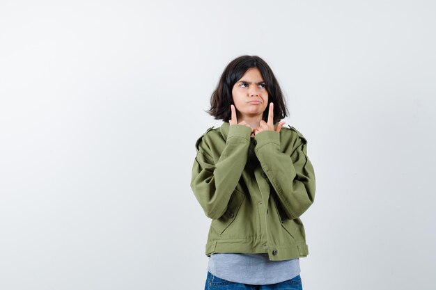 Chica joven apuntando hacia arriba con suéter gris, chaqueta caqui, pantalón de mezclilla y mirando pensativo. vista frontal.