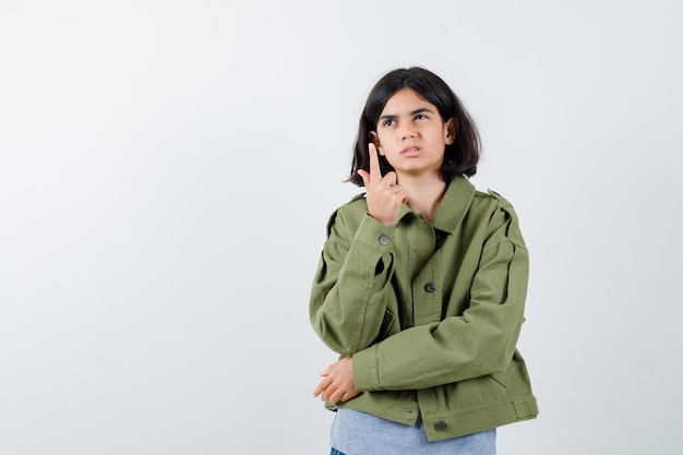 Chica joven apuntando hacia arriba con suéter gris, chaqueta caqui, pantalón de mezclilla y mirando pensativo. vista frontal.