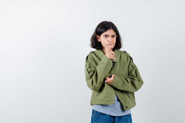 Chica joven apuntando con los dedos índices, guiñando un ojo en suéter gris, chaqueta caqui, pantalón de mezclilla y mirando seria. vista frontal.