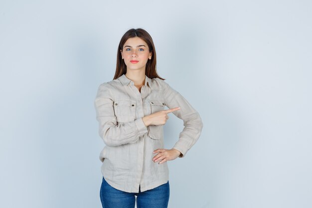 Chica joven apuntando a la derecha con el dedo índice, con la mano en la cadera en camisa beige, jeans y mirando serio, vista frontal.